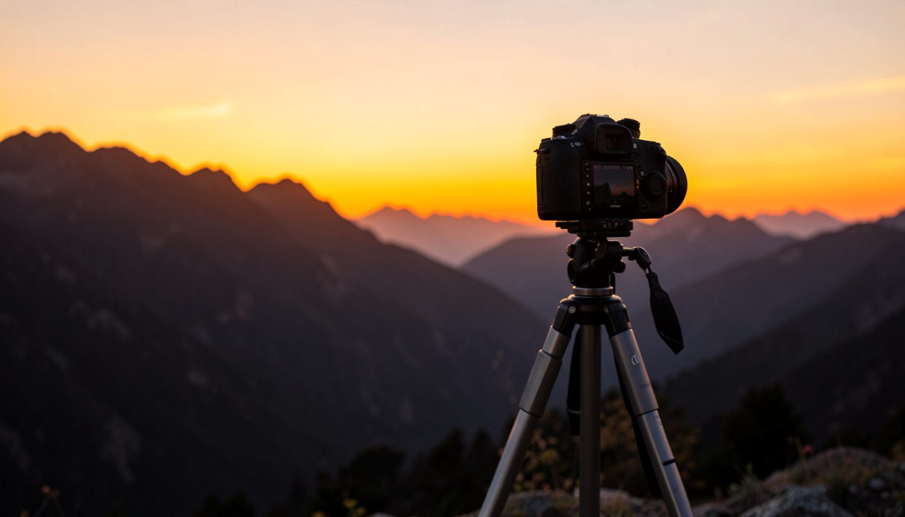 Cámara réflex sobre trípode junto a un paisaje montañoso al atardecer, tonos anaranjados y silueta de montañas